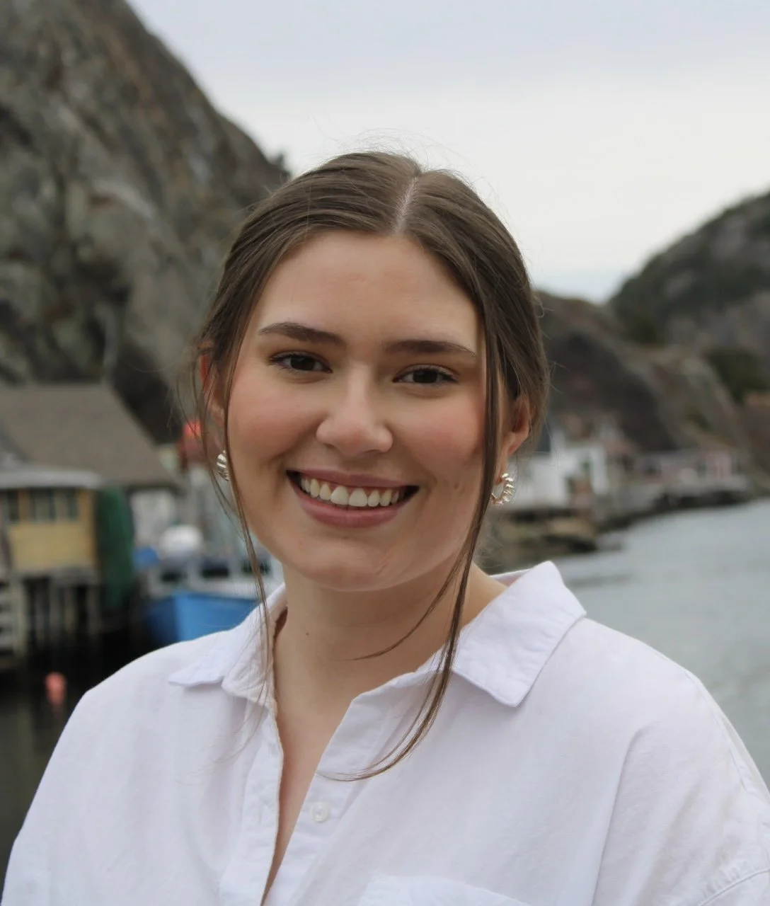 A smiling woman with brown hair in a white shirt outdoors near water and rocky cliffs.