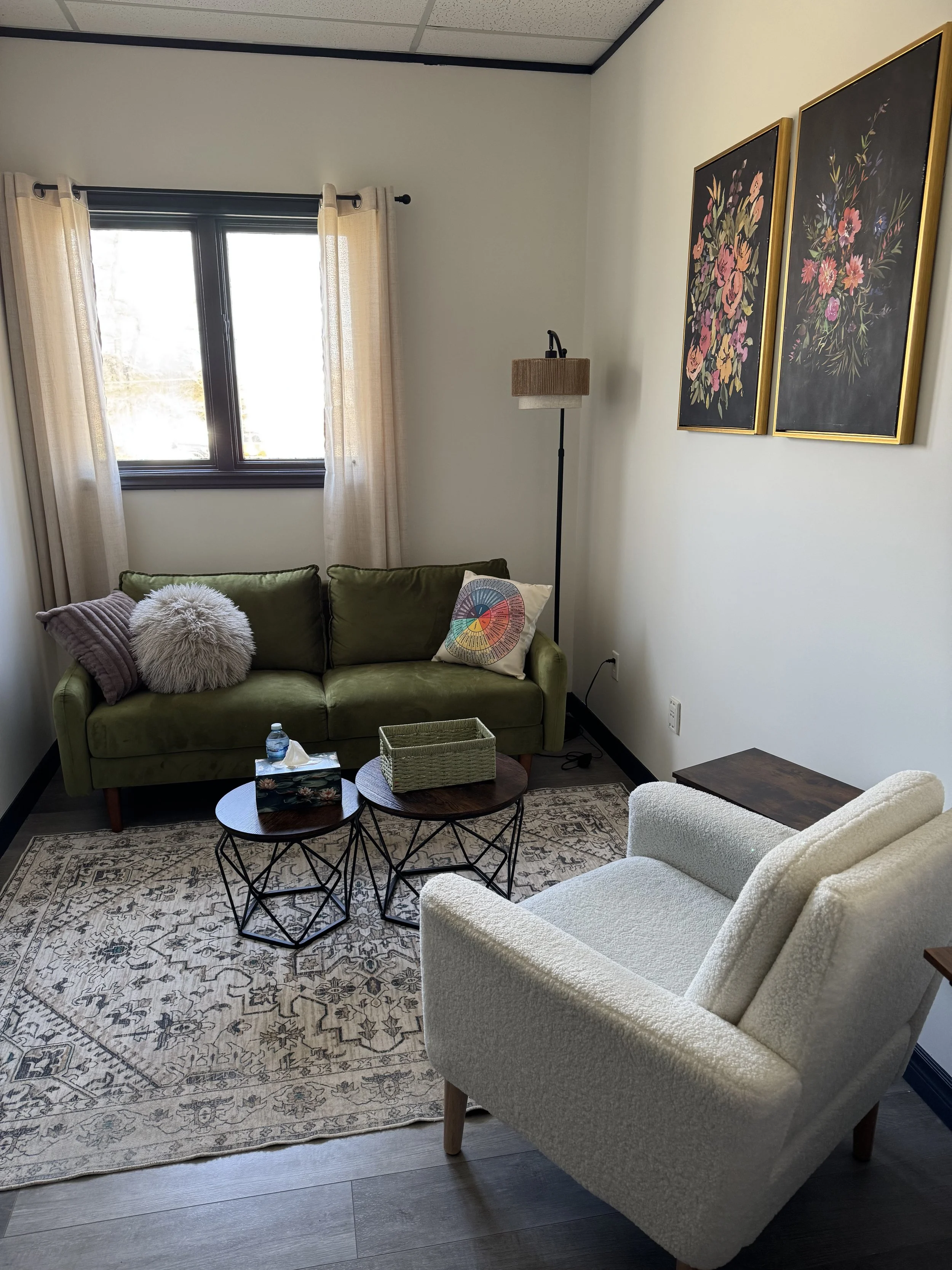 Living room with a white armchair, a sofa with green pillows, and a shaggy pillow. In front, there are three round black metal tables, with a tissue box, a water bottle, and a gift box on them. The room has a patterned rug on dark wood flooring, a window with cream curtains, wall art with floral designs, and a floor lamp.
