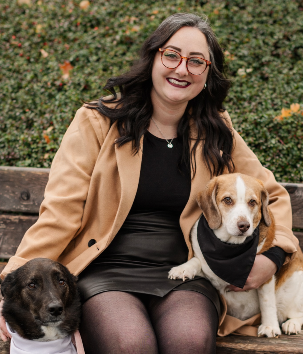 A woman with dark wavy hair, glasses, and dark lipstick sitting on a park bench, smiling, with two dogs, one sitting on her lap and the other on the ground beside her, all in fall outdoor setting with bushes in the background.