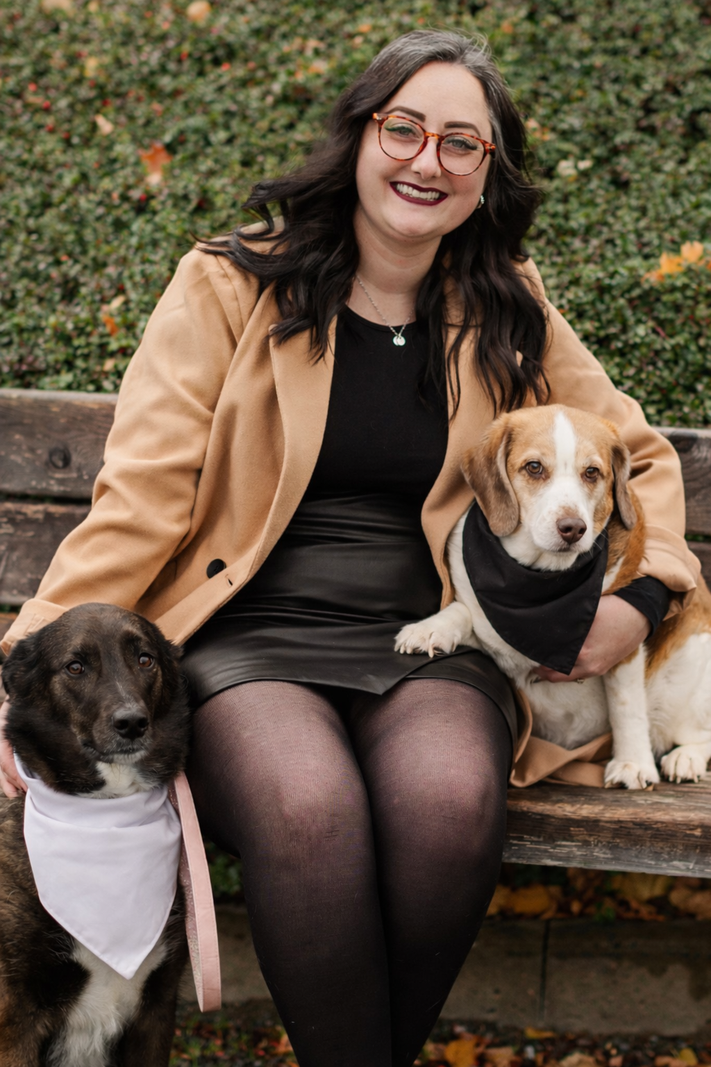 A woman with dark hair and glasses sitting on a park bench, smiling, with two dogs dressed in bandanas. One dog is sitting on her lap and the other is beside her.