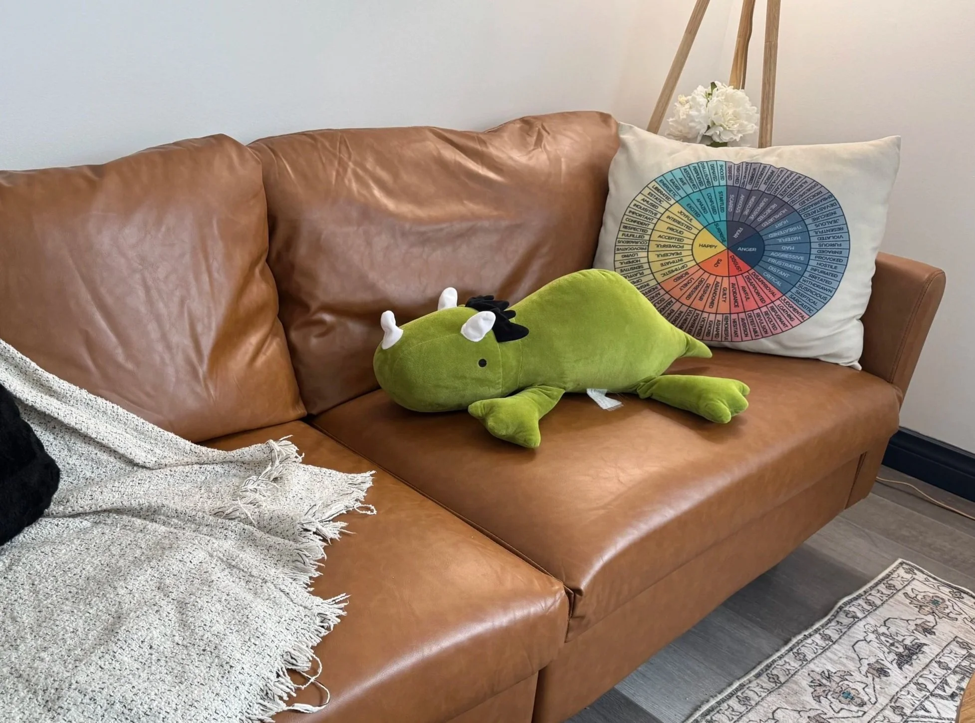A brown leather couch with a white textured throw blanket draped over the left armrest. On the right side of the couch, there is a white pillow with a circular design displaying various emotional words with colors in the background. A green plush toy resembling a lizard or crocodile with white horns and a black bow on its head is lying on the couch. In the background, a wooden ladder shelf holds a white flower arrangement. The floor is a dark wood, and a patterned rug is partially visible at the bottom of the image.