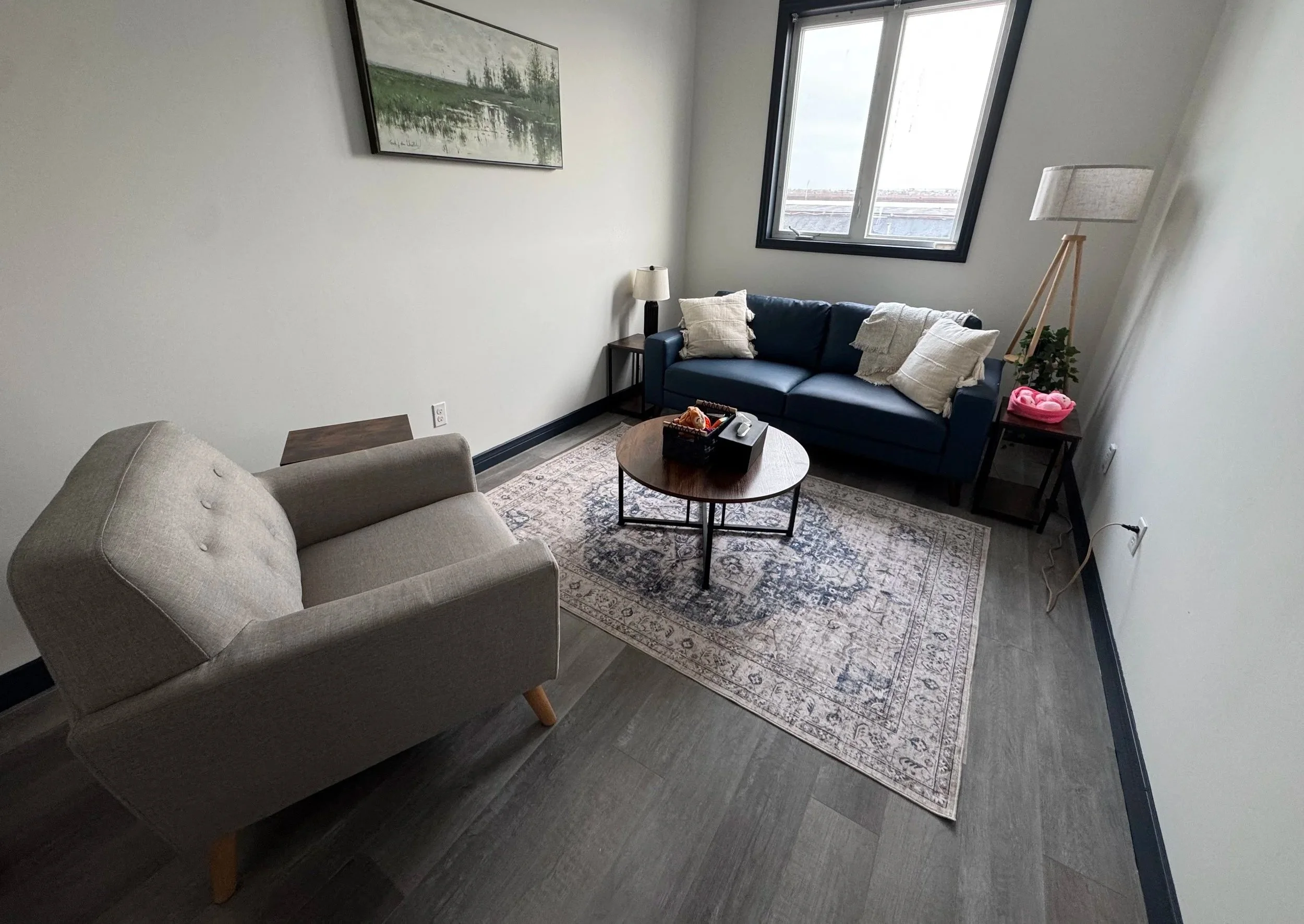 A living room featuring a beige armchair, dark blue sofa with pillows, a small wooden side table with a lamp, a round coffee table, patterned area rug, large window, and a wall painting of a landscape with trees and a river.