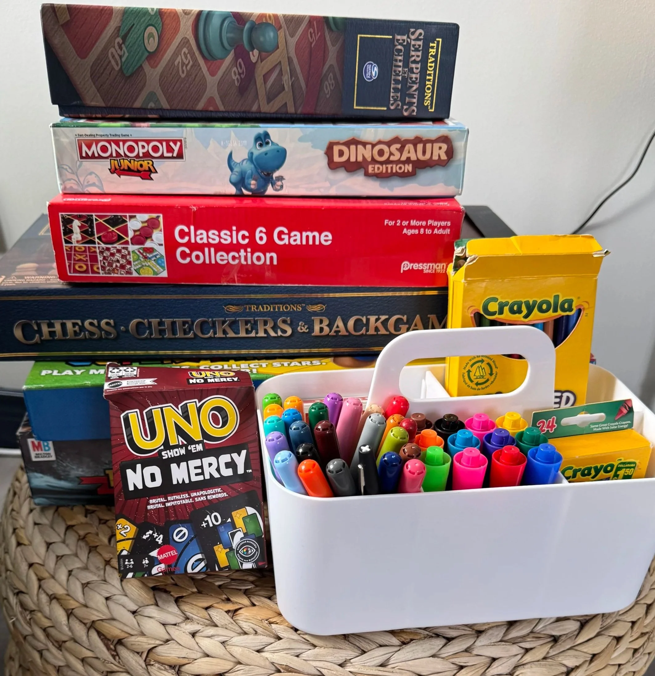 A stack of board games, including Monopoly Junior Dinosaur Edition, Chess, Checkers, Backgammon, and Uno, with colorful markers in a white container.