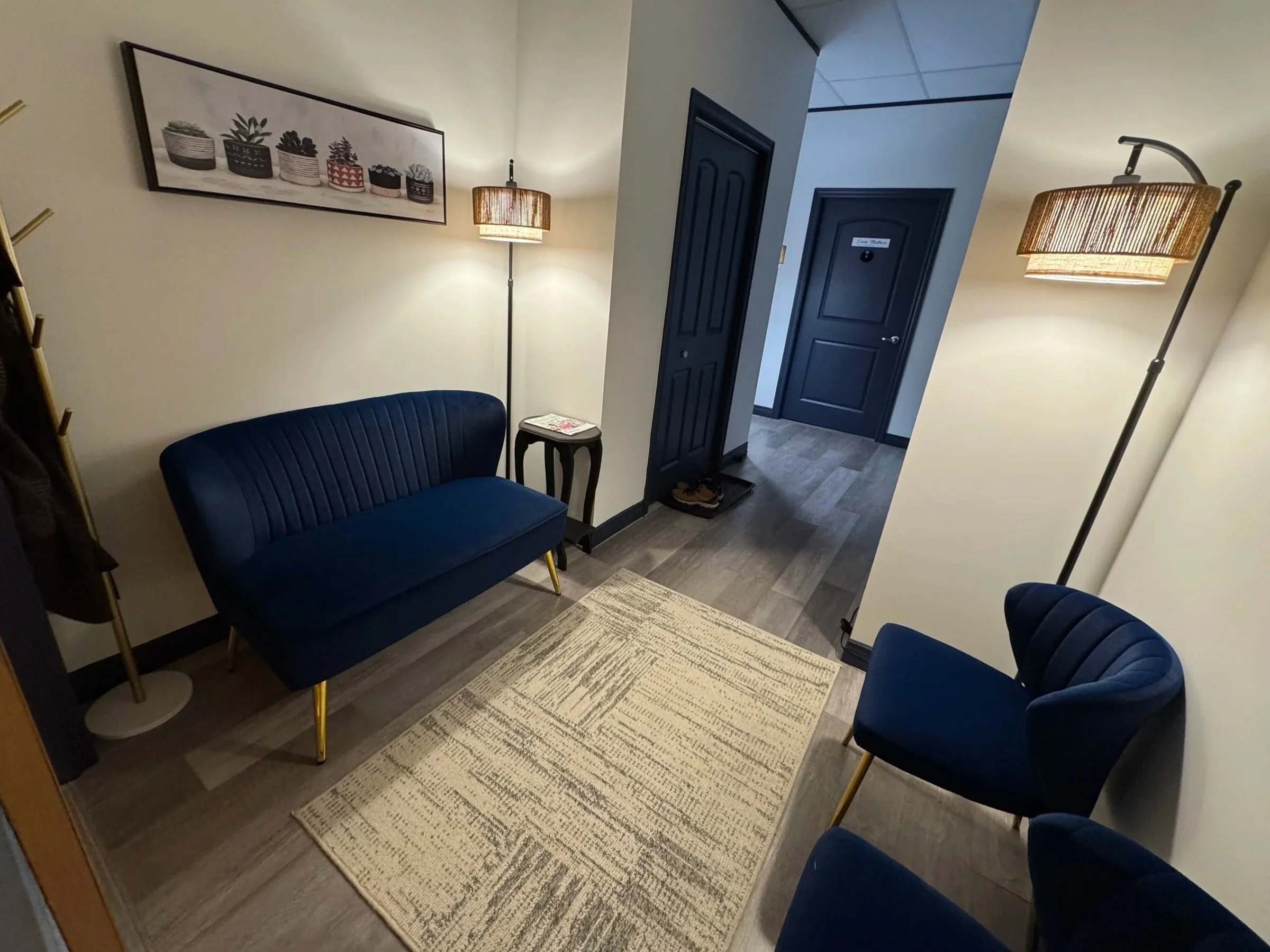 A lobby or waiting area with a blue velvet couch, matching blue armchair, two window lamps with woven shades, a framed art print of potted plants, and a small table with magazines. There are two closed black doors, one labeled 'Case 1'. A beige rug is on the wood-look floor.