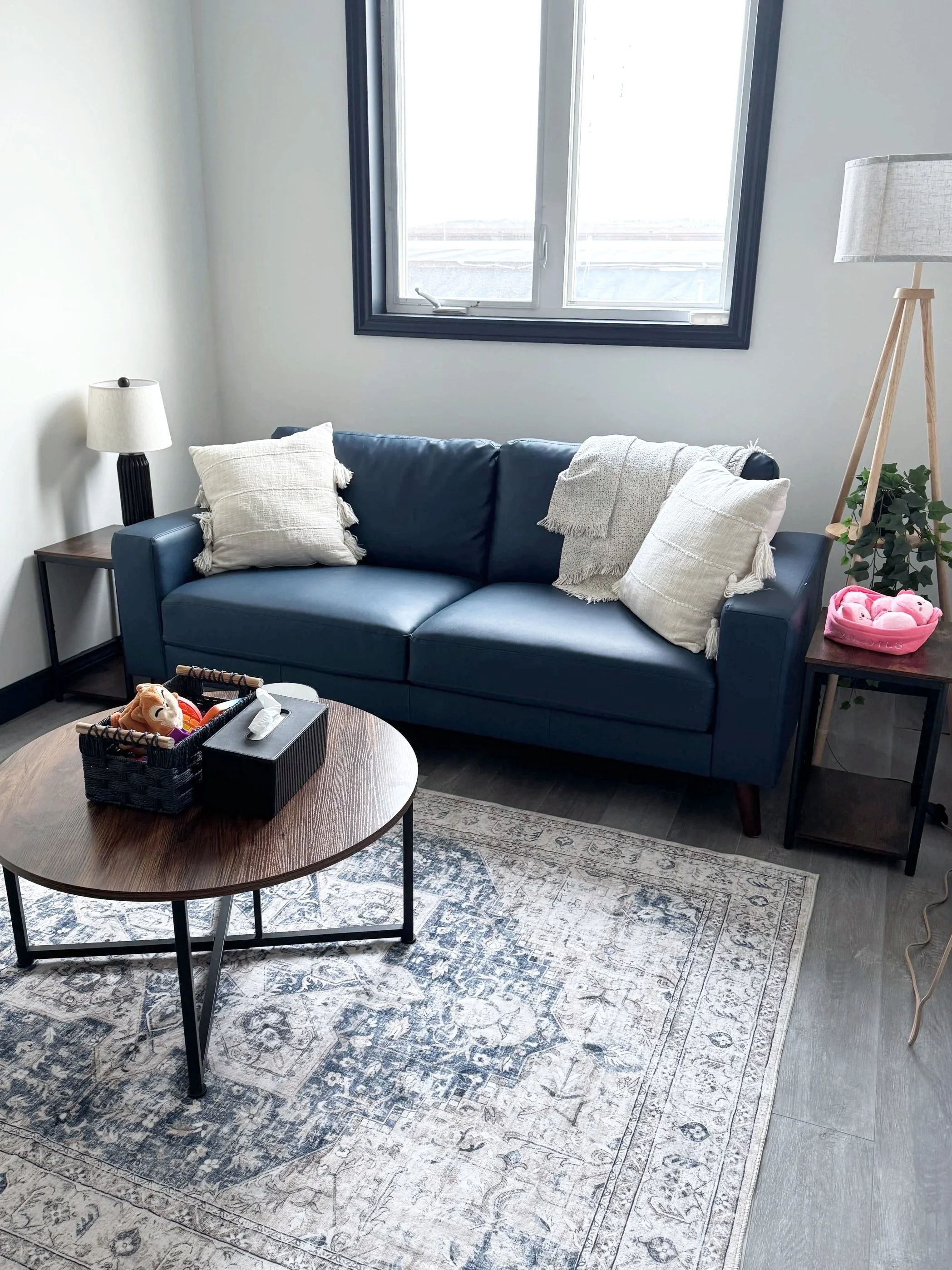 A living room with a blue couch, white pillows, a beige blanket, a wooden side table with a plant and pink plush toys, a black side table with a lamp, a round wooden coffee table with toys, and a patterned rug on a gray wooden floor, with a window