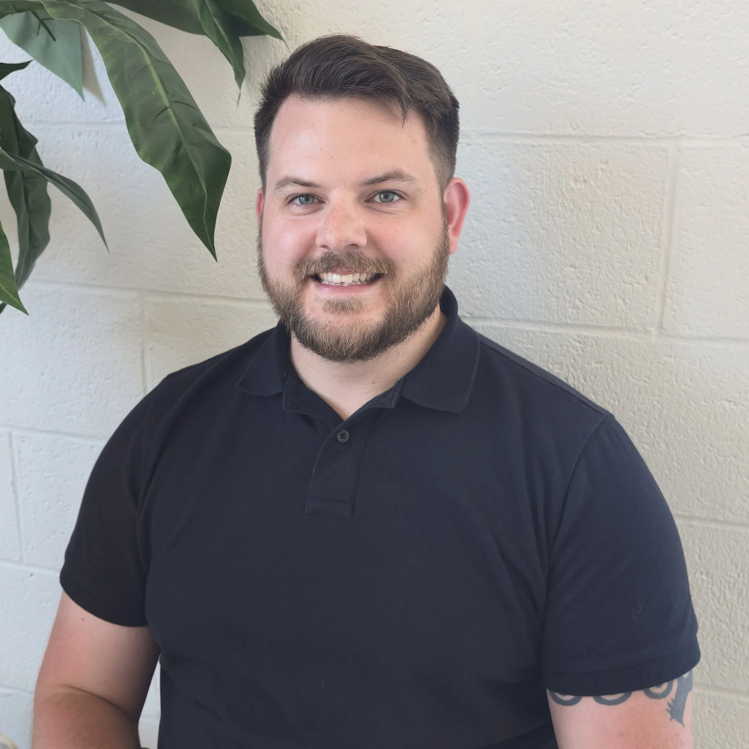 Close-up of a smiling man with short brown hair, a beard, wearing a black shirt, standing in front of a white brick wall and a large green plant.