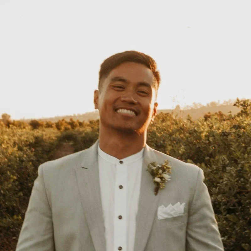 A smiling man in a light gray suit with a white shirt, standing outdoors during sunset in a field of bushes, with a small flower pin on his lapel.