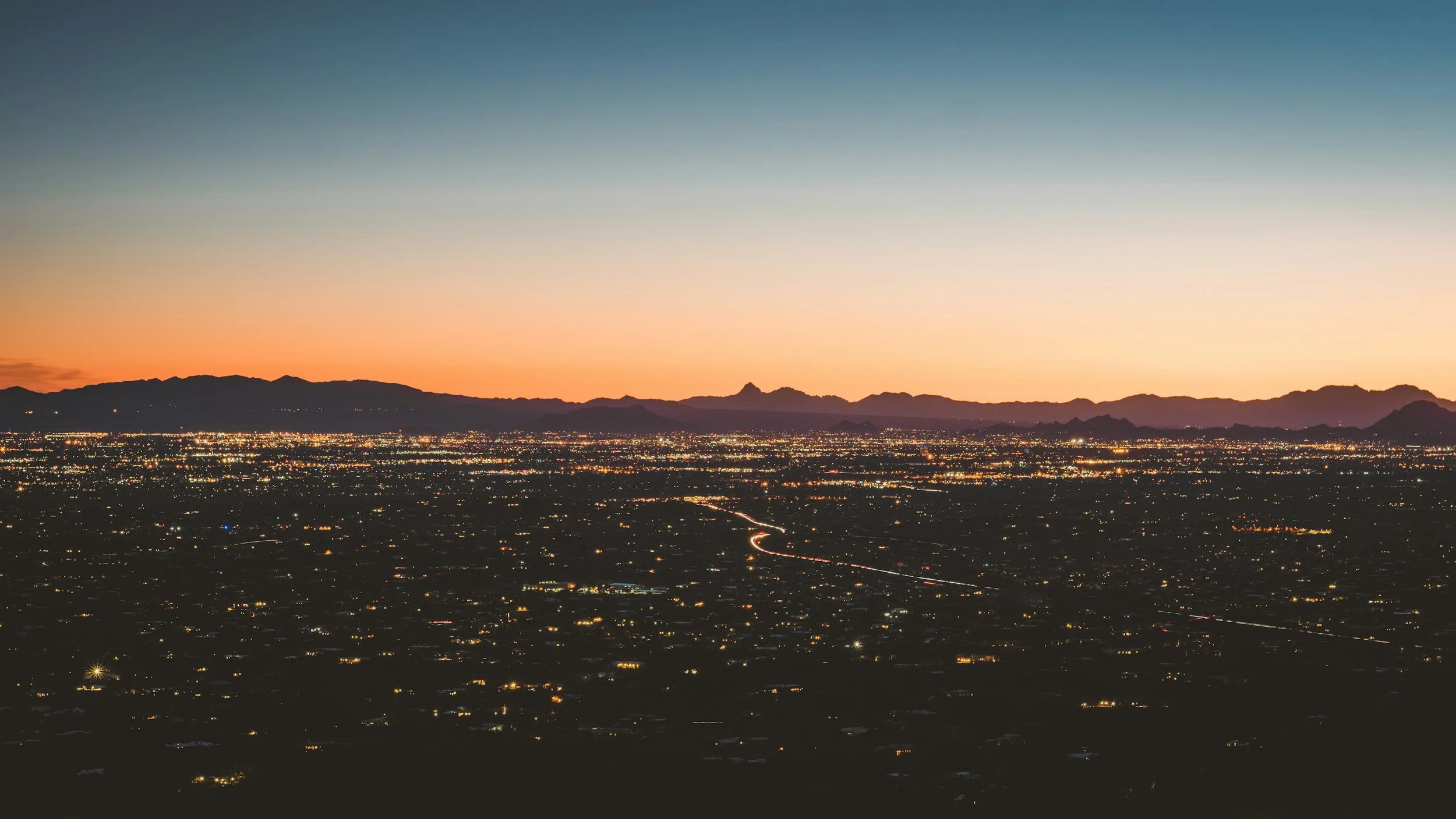 A view of a Tucson Arizona at dusk with a sunset sky and distant mountains, city lights illuminating the landscape.