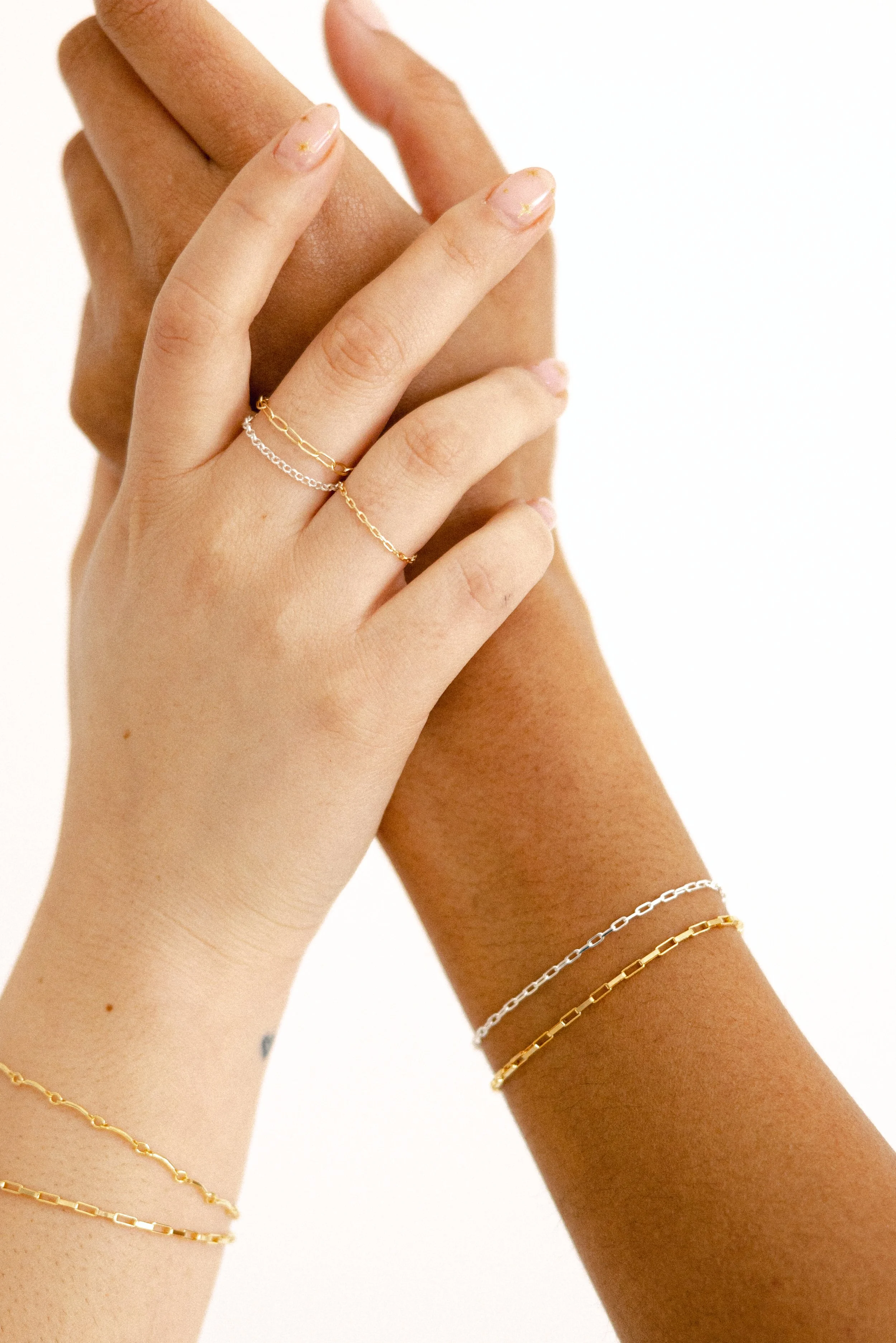 Close-up of two hands with different skin tones, wearing gold and silver chain bracelets and rings, with a plain white background.