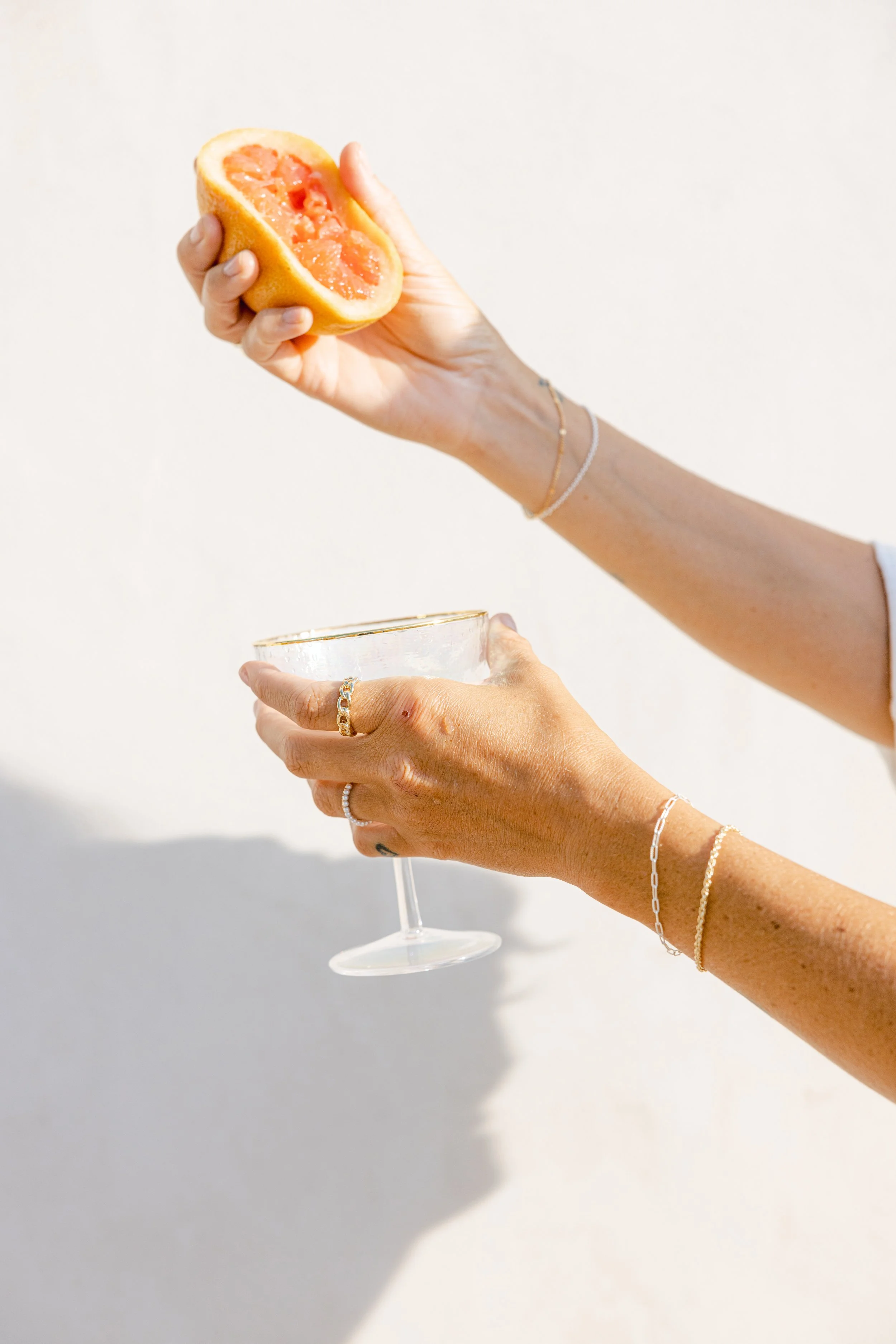 A person's hand holding a half grapefruit above a glass of champagne, with jewelry on the hand and wrist, against a plain white wall background.