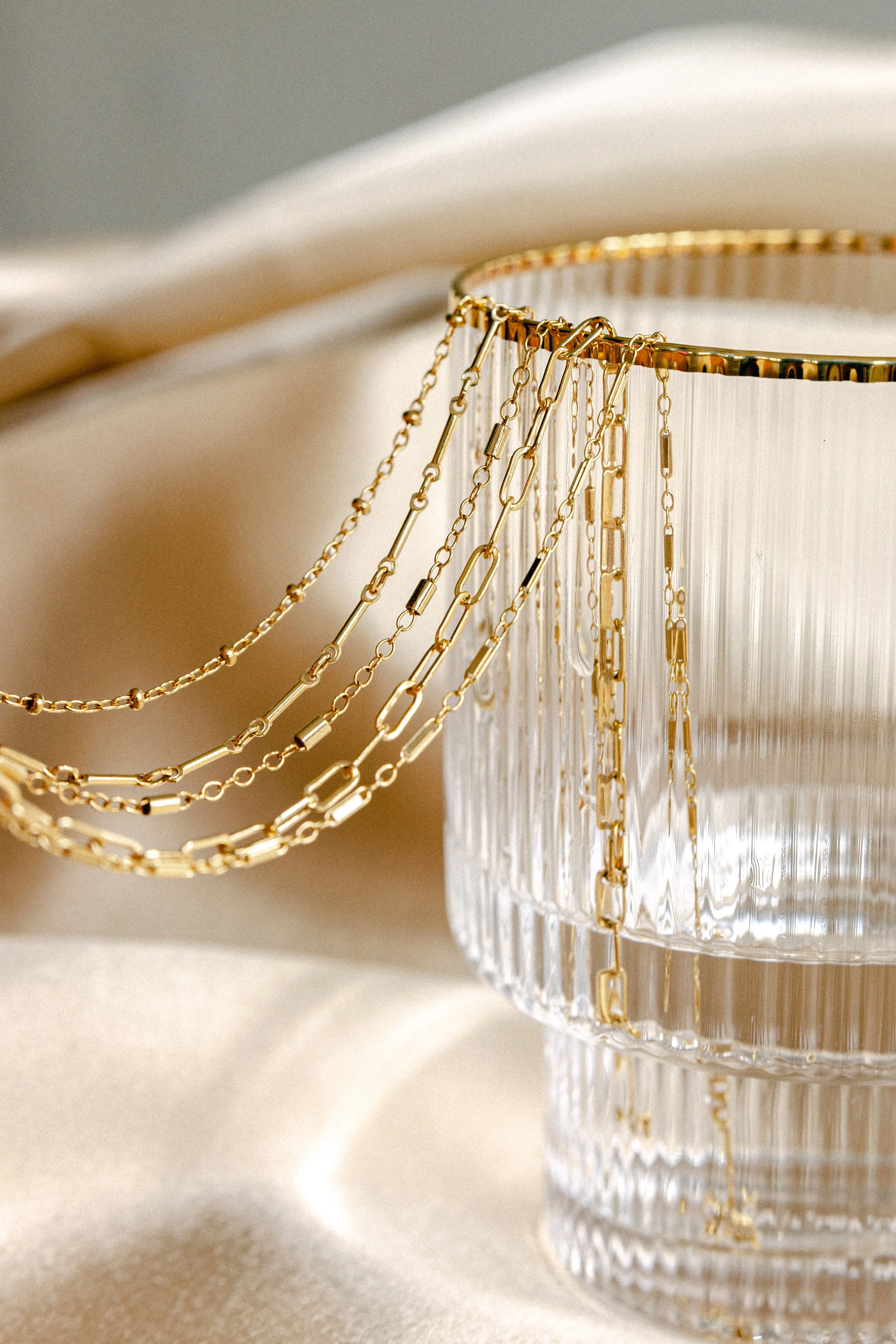 Close-up of gold chains draped over a clear, textured glass container, with a soft beige background.