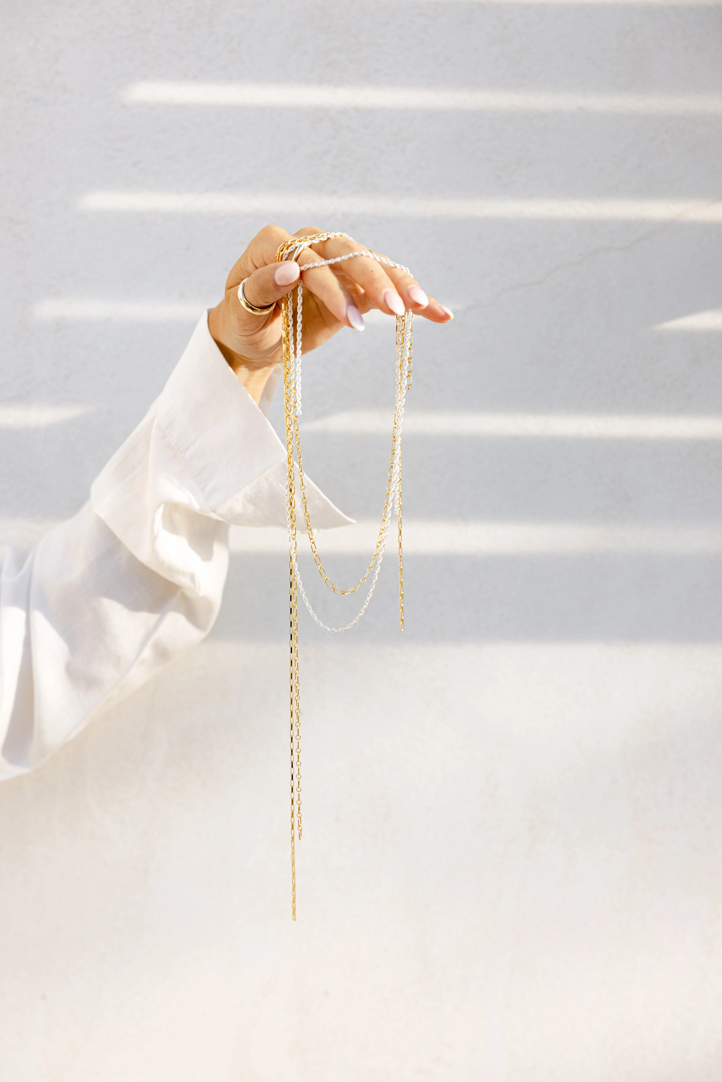 A hand with painted nails holding multiple gold and silver necklaces against a light background with horizontal shadows.