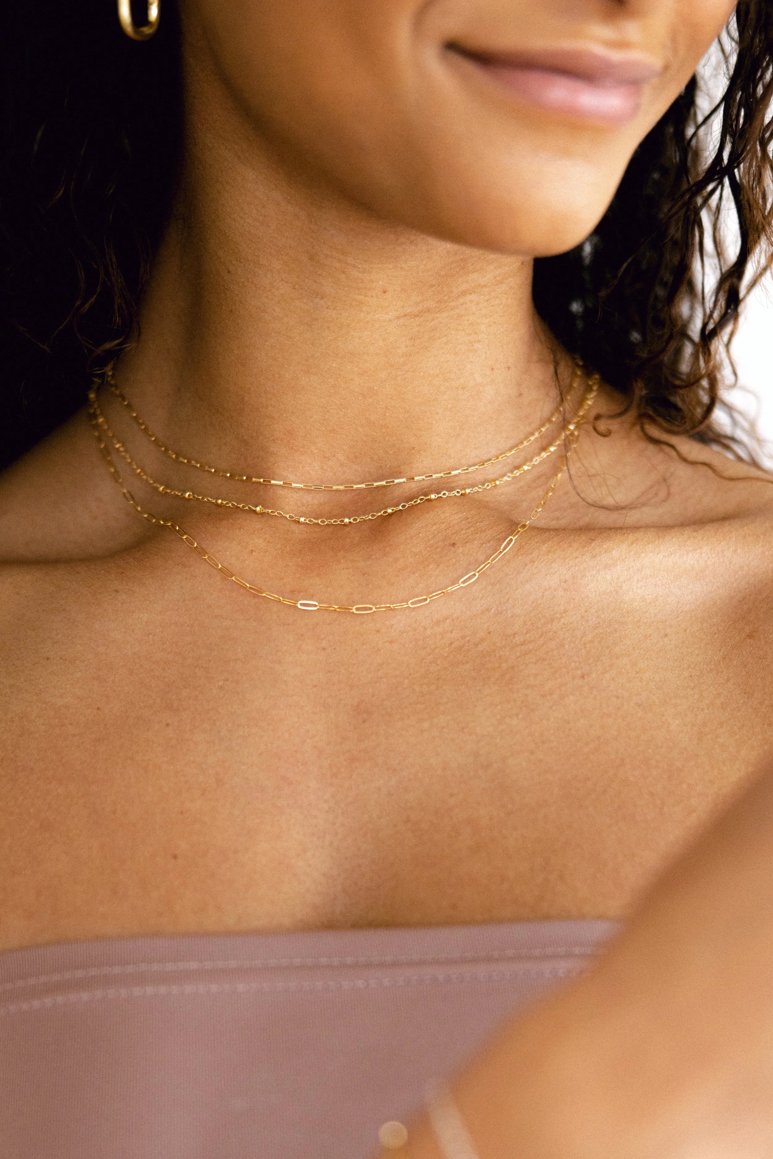 Close-up of a woman's neck, wearing layered gold necklaces, partially visible face, and curly hair.
