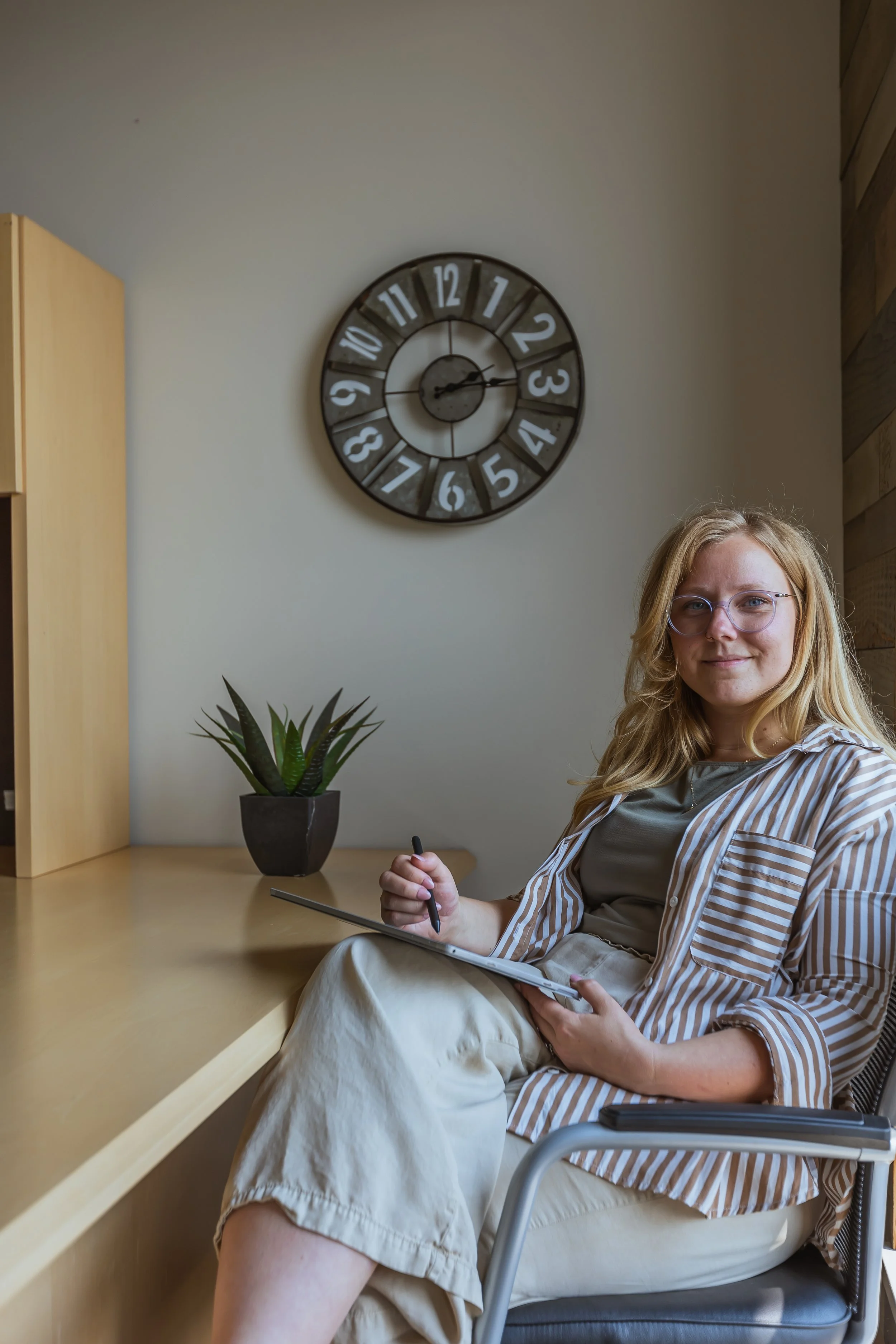 A woman with long blonde hair and glasses sitting at a desk, holding a pen and paper, in a room with a wall clock showing approximately 2:07.