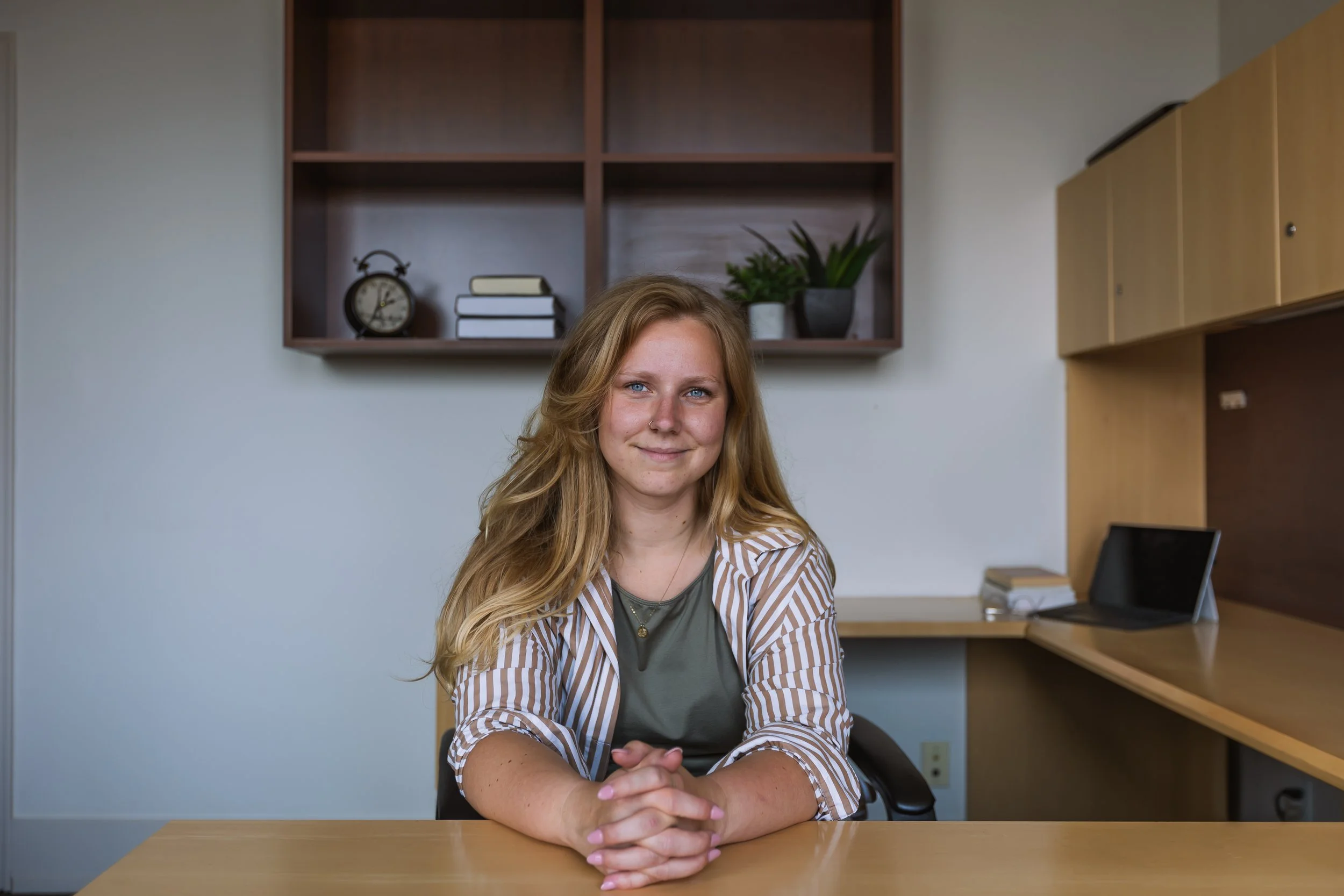 A young woman with long blonde hair, blue eyes, and light skin sitting at a desk in an office. She is smiling softly with her hands clasped on the desk and wears a green top and a striped blazer. The background shows office shelves with books, a clock, and a potted plant.