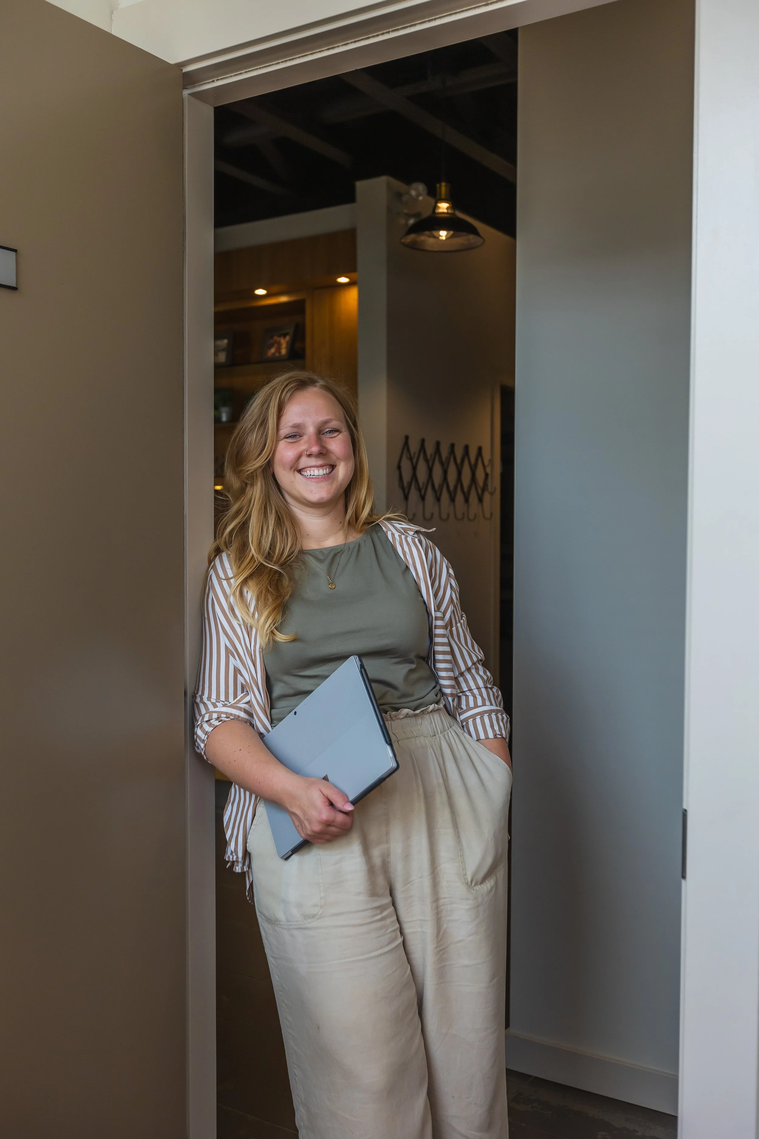 Smiling woman with blonde hair holding a closed laptop, standing in a room with gray and wooden walls.