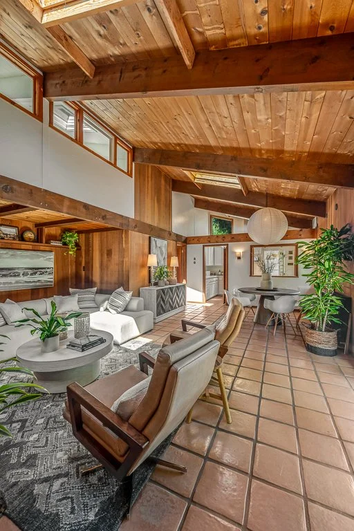 Interior of a living room with wooden ceiling beams, terracotta tile flooring, a white sofa with patterned pillows, a round coffee table with plants, armchairs, a dining table with chairs, and large windows allowing natural light.