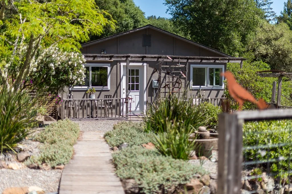A small house with a deck, surrounded by lush greenery and plants, with a pathway leading to the front entrance.