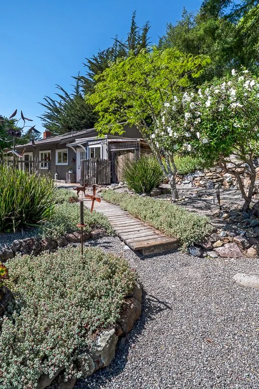 A small wooden footbridge over a gravel garden path leading to a house, surrounded by lush green shrubs and trees under a bright blue sky.