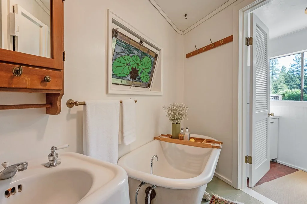 Bathroom with a vintage clawfoot bathtub holding a tray with a flower vase and toiletries, a white sink, a small stained glass window, towel rack with towels, wooden hooks, and a louvered door leading to a sunlit room with trees outside.