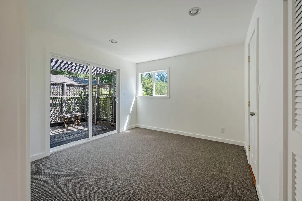 Empty room with white walls and carpeted floor, sliding glass door leading to a small outdoor patio, small window, and a closet with louvered doors.