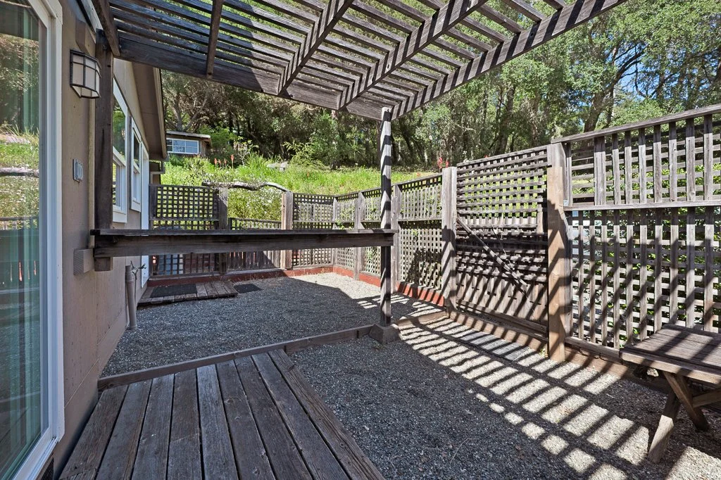 Backyard patio with wooden lattice and pergola casting striped shadows, gravel ground, and a wooden deck.