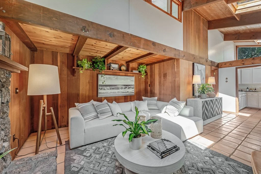 Living room with wood-paneled walls, white sectional sofa with patterned pillows, potted plants, and large windows allowing natural light. A lamp beside the sofa and a framed picture on the wall enhance the cozy, modern rustic decor.