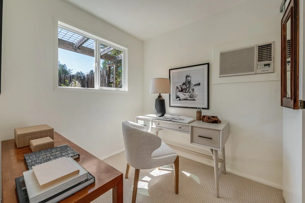 A small home office with a window, a white desk, a white chair, a lamp, a framed photo, and a wall air conditioner