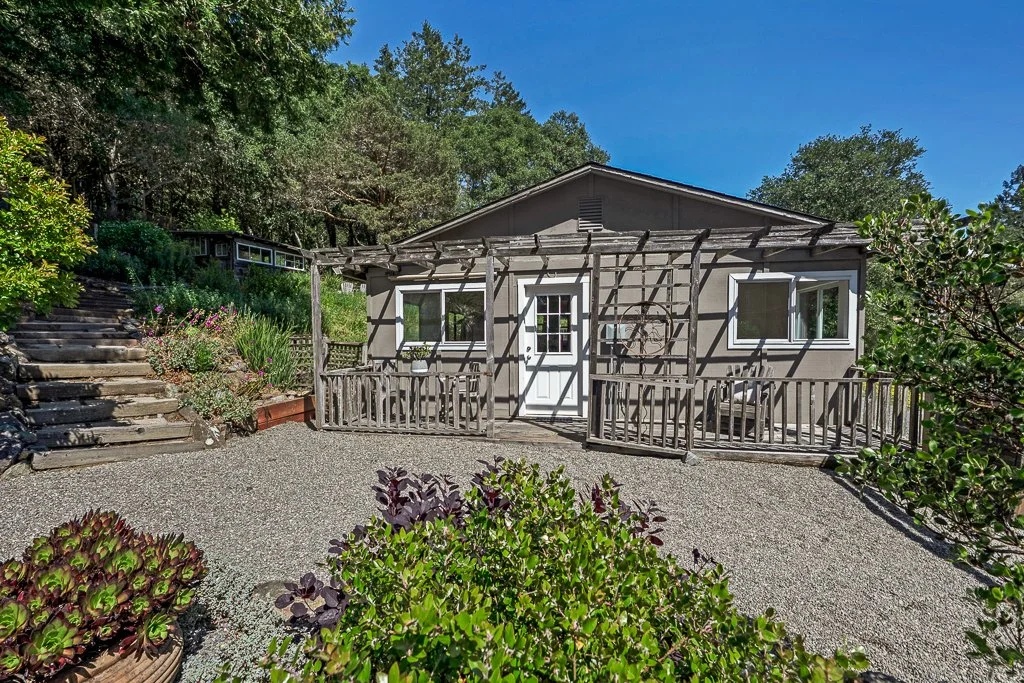 Small house with a wooden deck and trellis, surrounded by plants and trees under a blue sky.