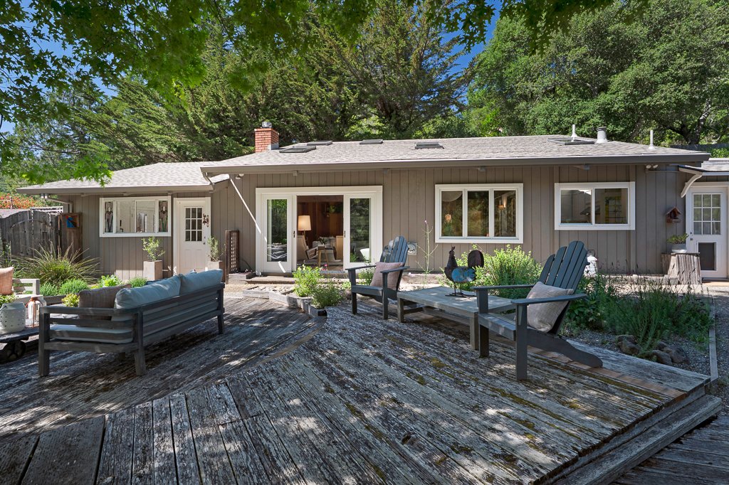 Backyard patio with wooden deck, outdoor seating, and sliding glass door leading into house, surrounded by green trees