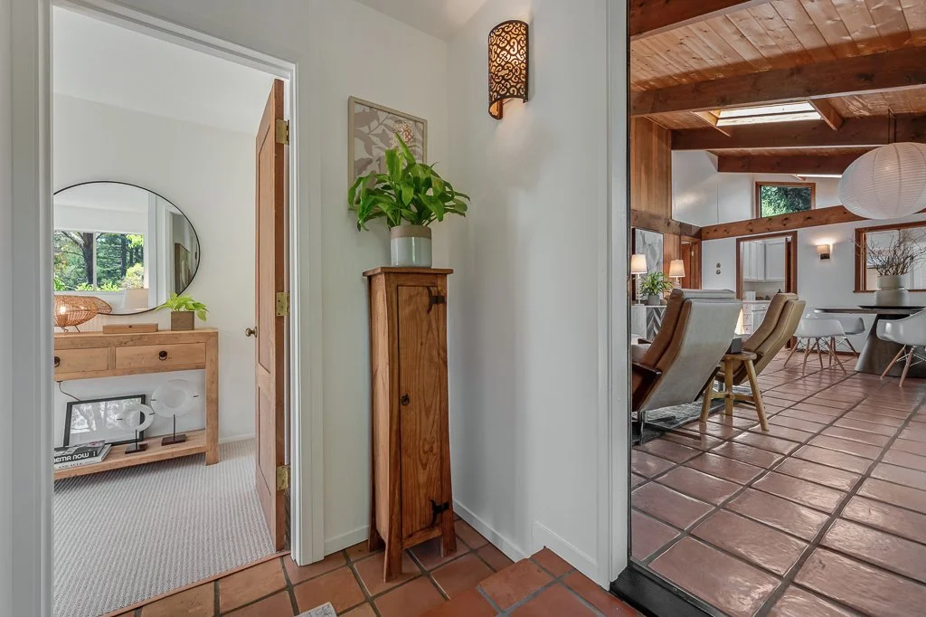 View of a hallway with white walls, a tall wooden cabinet, a potted plant, and a wall-mounted light fixture. Through an open door, a room with a mirror, a wooden table, and a window is visible. To the right, an open living and dining area with wooden