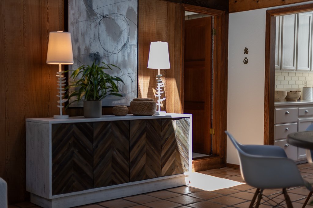 Living room with a white sideboard, two lamps, a potted plant, and decorative bowls, next to a doorway leading to a kitchen area with white cabinets and neutral-colored bowls.