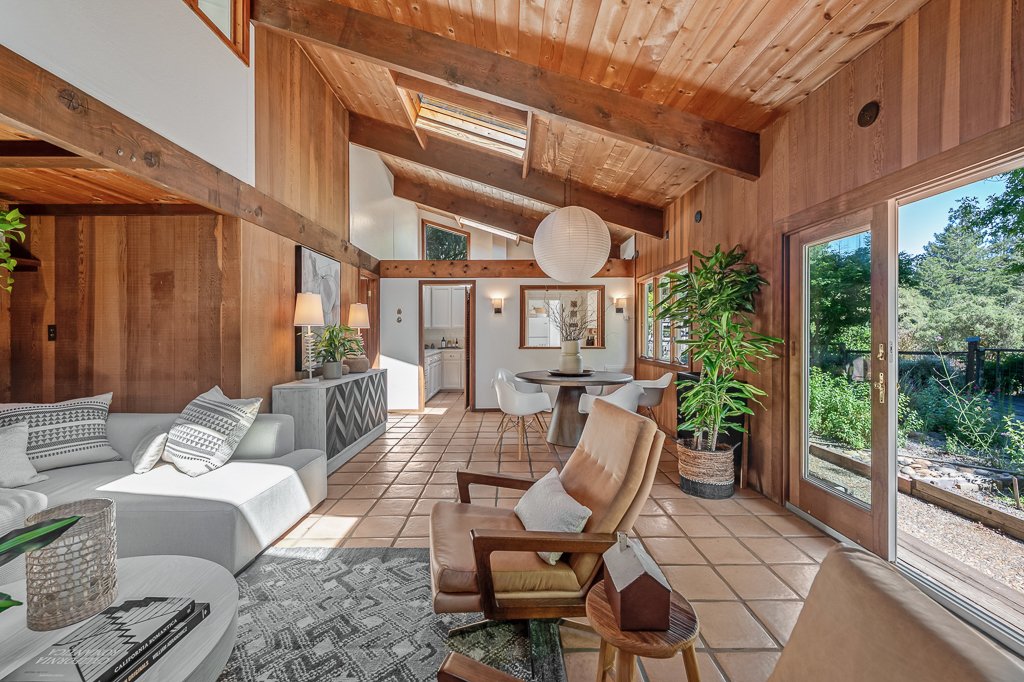 Living room with wood-paneled walls, large windows, and a sliding glass door leading outside, featuring a beige couch, armchairs, a round dining table, potted plants, and skylights.