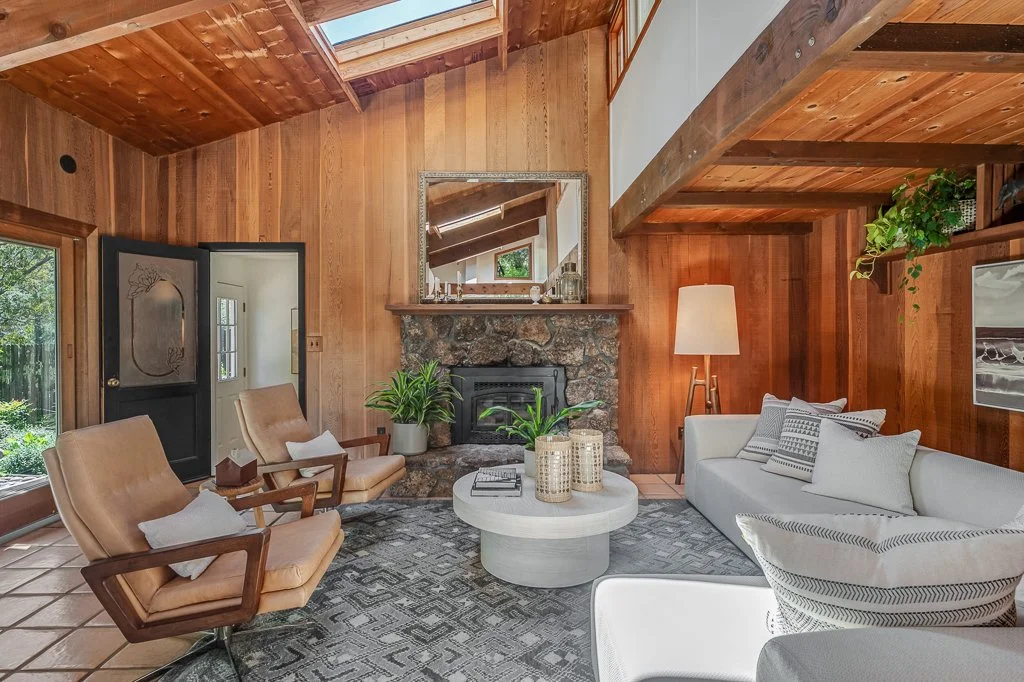 Living room with wood-paneled walls and ceiling, stone fireplace, large window, beige armchairs, white sofa with pillows, coffee table with decorative items, potted plants, floor lamp, and a mirror above the fireplace.