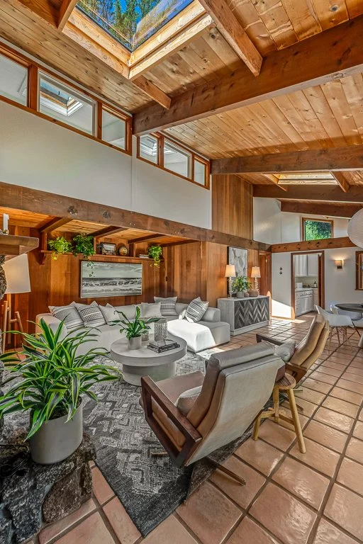 Living room with wooden ceiling beams and skylights, white sectional sofa, gray armchairs, potted plants, and rustic decor.
