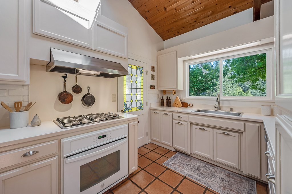 Kitchen with white cabinets, a gas stove with an oven, a range hood, a double sink under a large window, and hanging pots. Decor includes bottles, a cutting board, and window with stained glass design. The ceiling is wood-paneled.