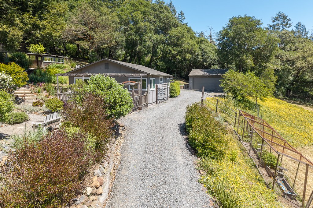 Gravel driveway leading to a house and a detached garage, surrounded by greenery and trees, on a sunny day.