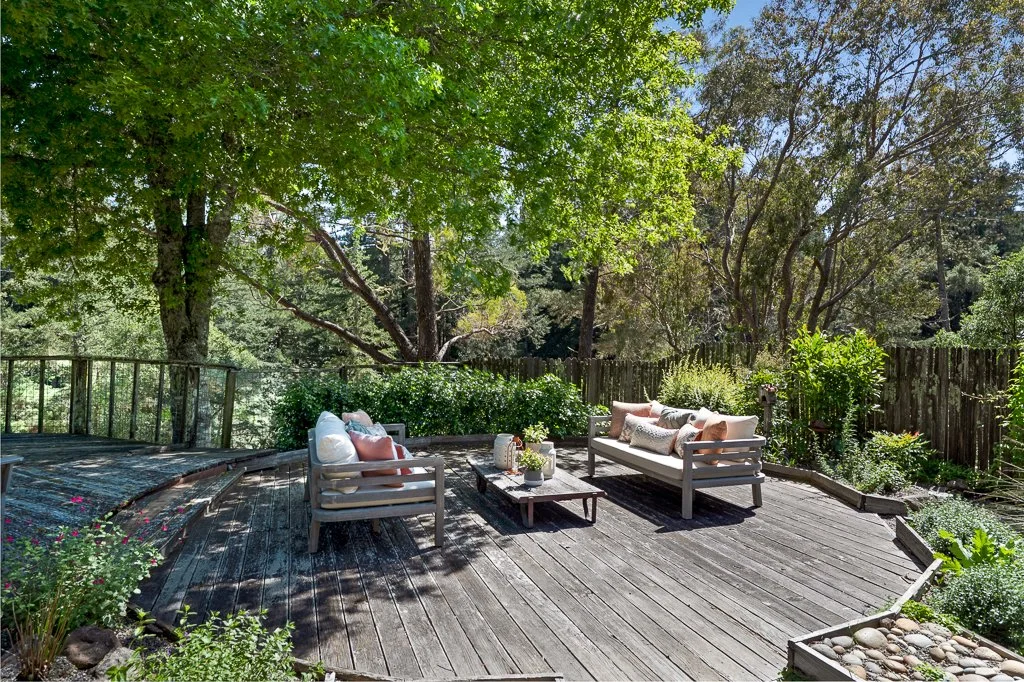 Outdoor wooden deck with outdoor furniture including two cushioned sofas and a coffee table, surrounded by lush greenery and trees.