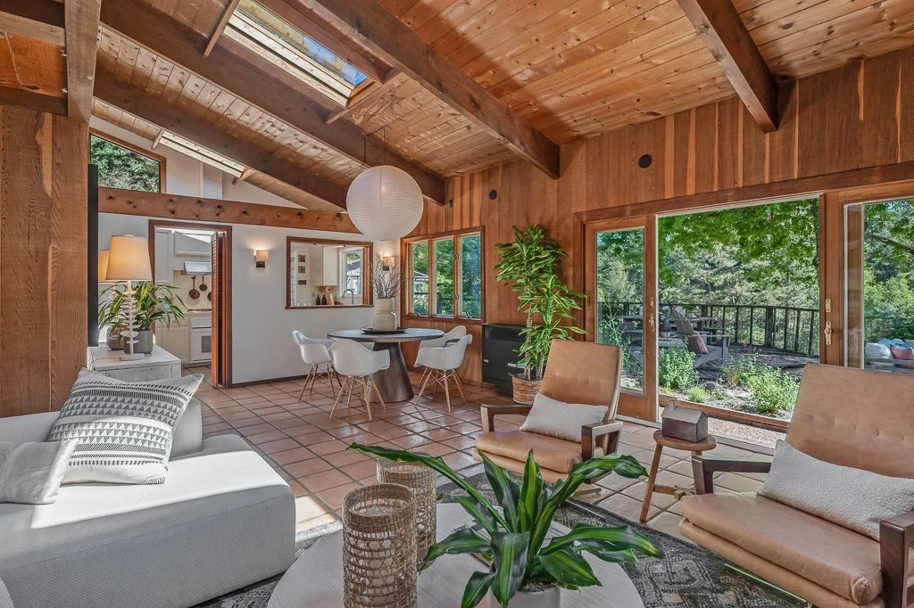 Living room with wood-paneled walls and ceiling, large sliding glass door leading to an outdoor deck, and various chairs and plants inside.