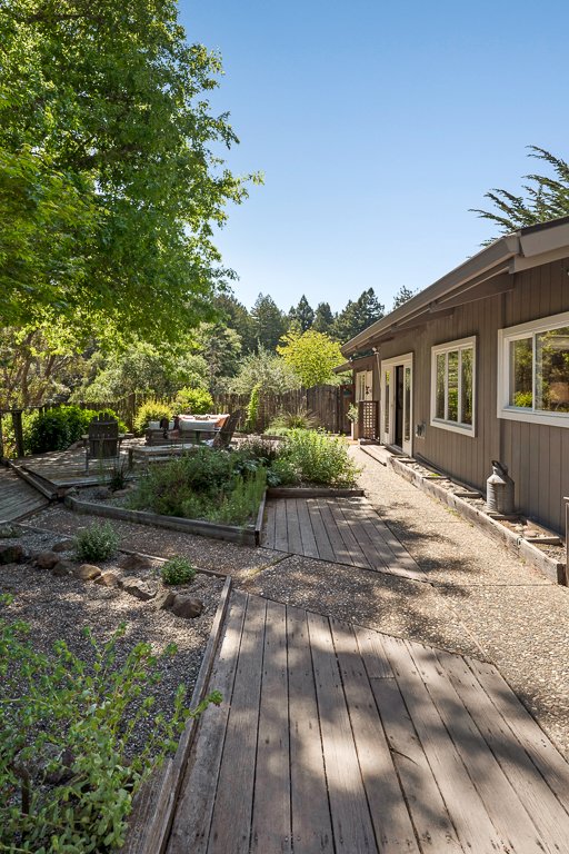 Backyard garden with wooden pathway, trees, shrubs, outdoor furniture, and a house with large windows under a clear blue sky.