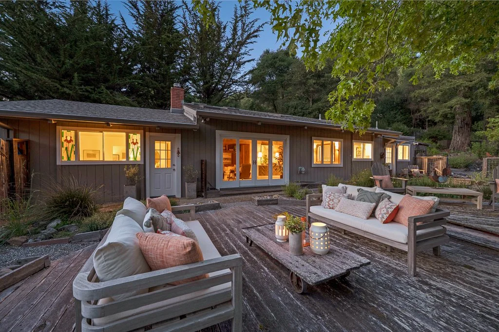 Backyard patio at dusk with wooden flooring, outdoor seating with cushions and pillows, lanterns, and a gray house with lit windows surrounded by trees.