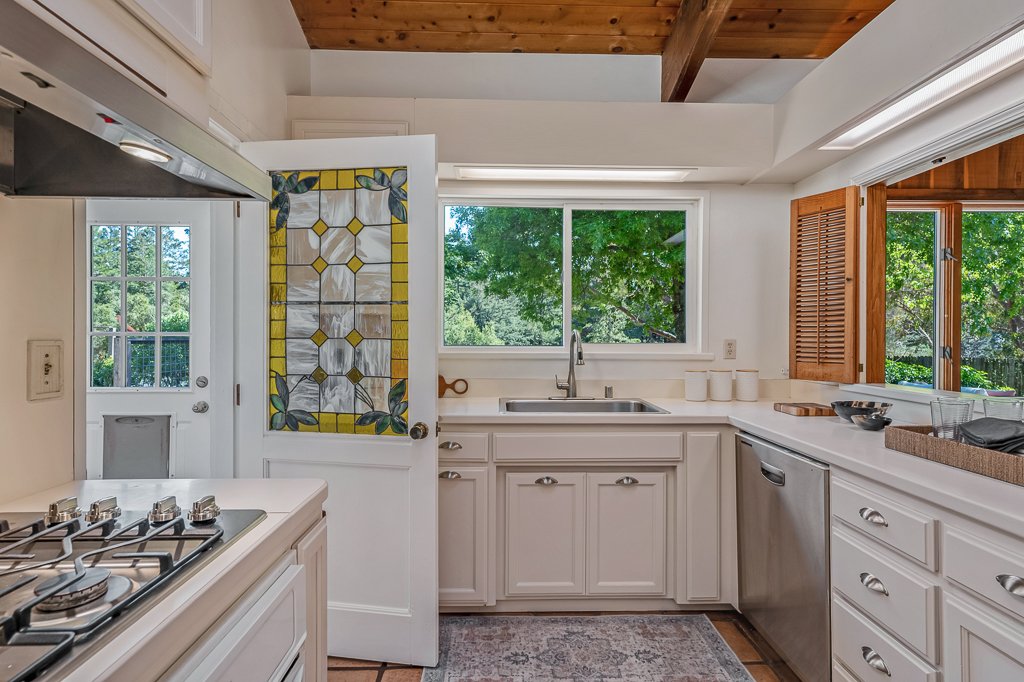 Kitchen with white cabinets, a window over the sink, a stove on the left, and a dishwasher on the right. Green trees are visible outside through the windows and a door.