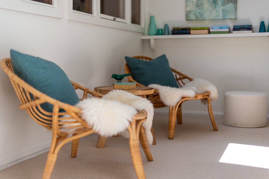 Cozy sitting area with rattan chairs, teal pillows, sheepskin throws, a small side table with a decorative bird, and a white ottoman in a bright room with books and artwork on the wall.
