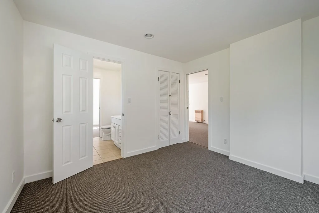 Empty room with white walls, gray carpet, and two open doors leading to bathroom and another room, with a closet in the wall.