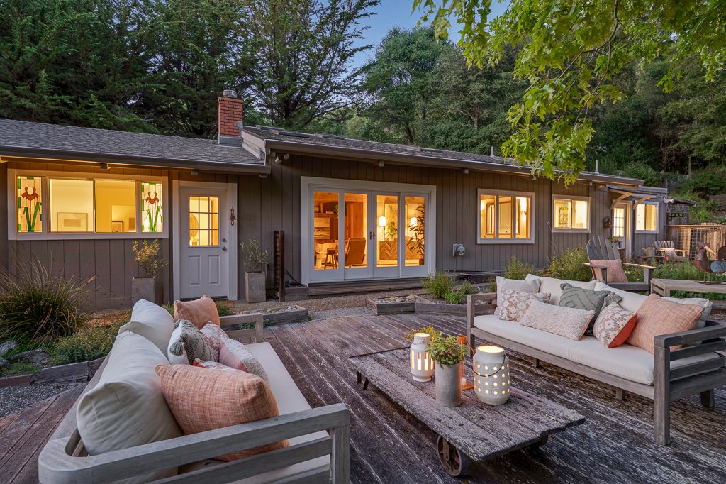 Backyard patio of a house with outdoor furniture, lanterns, and decorative pillows, with house interior lit up through large windows and sliding doors, surrounded by trees.