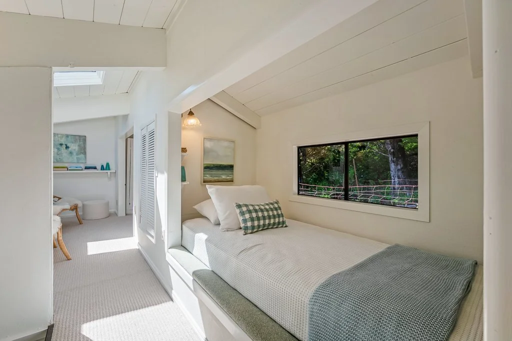 A cozy bedroom with white walls, a skylight, a window overlooking greenery, and a bed with white bedding and pillows.
