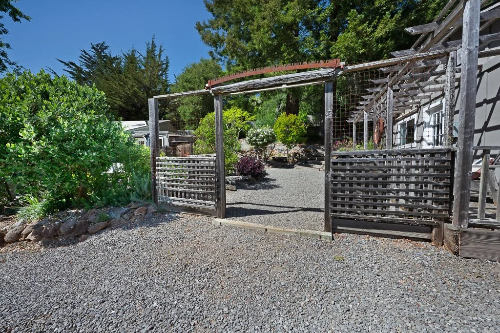 A garden area enclosed by a wooden fence with a gate, featuring gravel ground, various shrubs, and trees under a clear blue sky.