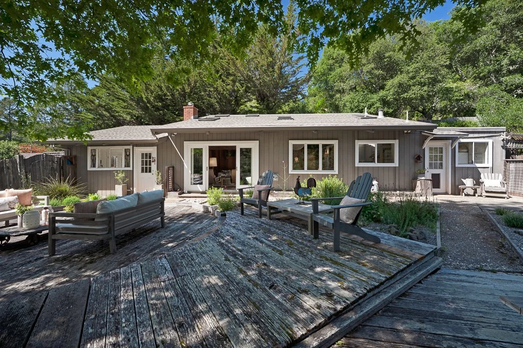 Front view of a house with a wooden deck, outdoor furniture, and lush trees providing shade.