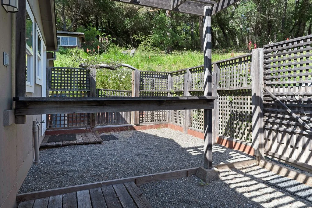 Backyard enclosed with wooden fencing, gravel ground, partial deck, and shaded area with trees in the background.