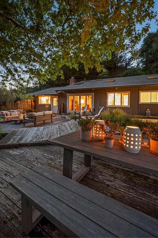 A cozy backyard outdoor patio area at dusk with wooden furniture, potted plants, and warm outdoor lighting, surrounded by trees and a house with lit windows.